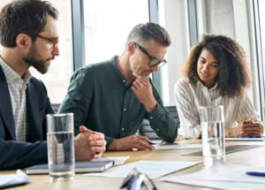 Three people are sitting at a conference table, engaged in discussion, with documents and water glasses in a modern glass-walled office setting.