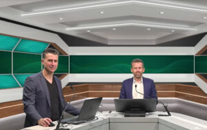 Two people in a modern studio with green, wood, and white decor, standing at podiums with laptops and microphones.