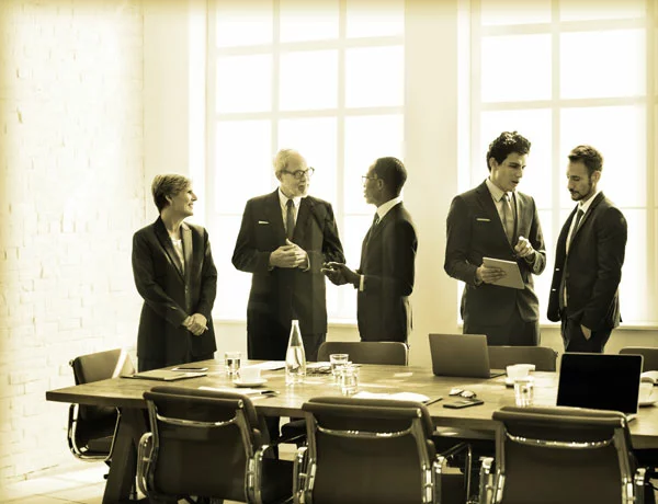 Five people in business attire are engaged in discussion around a conference table, with laptops and documents, in a brightly lit room.