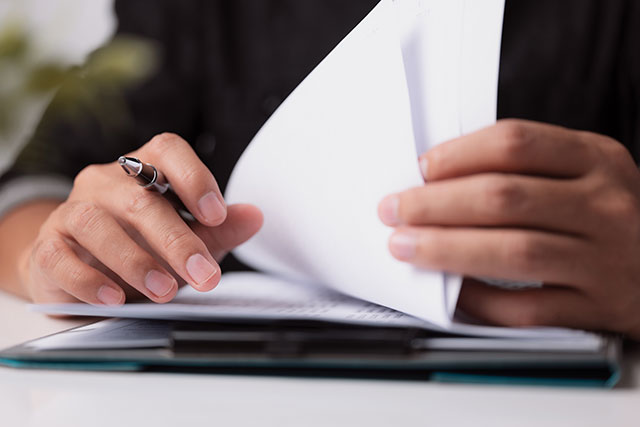 A person reviews documents on a clipboard, holding a pen. Close-up of hands flipping through pages in a focused, professional setting.