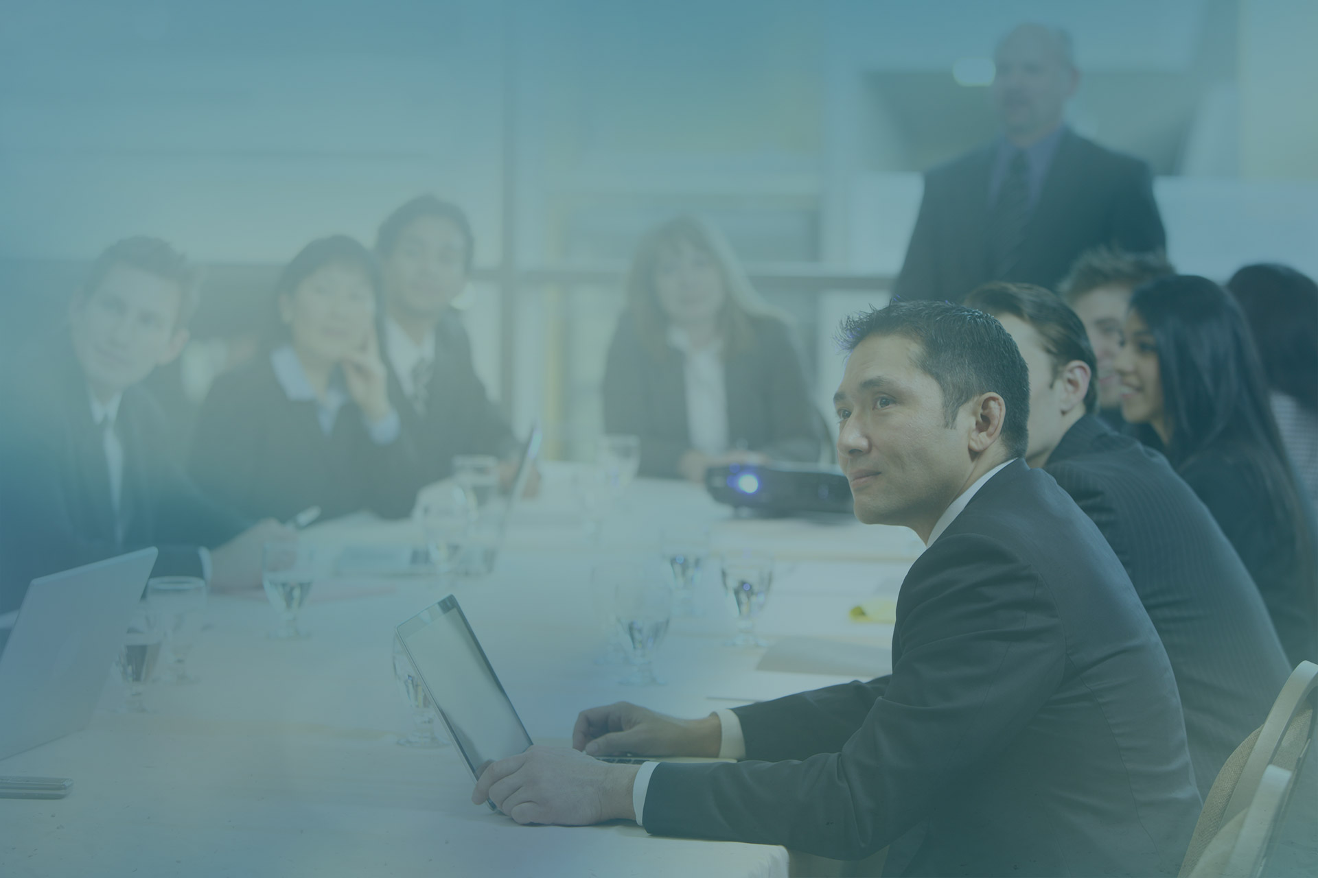 A group of individuals in formal attire sit around a conference table in a modern office setting, engaged in a meeting or discussion.