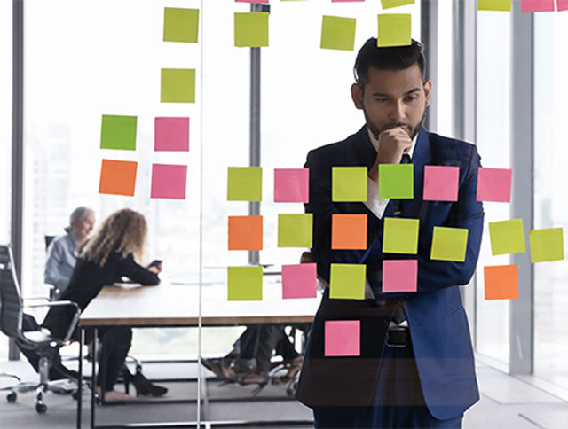 A person in an office is contemplating sticky notes on a glass wall. Two people converse in the background by a window.