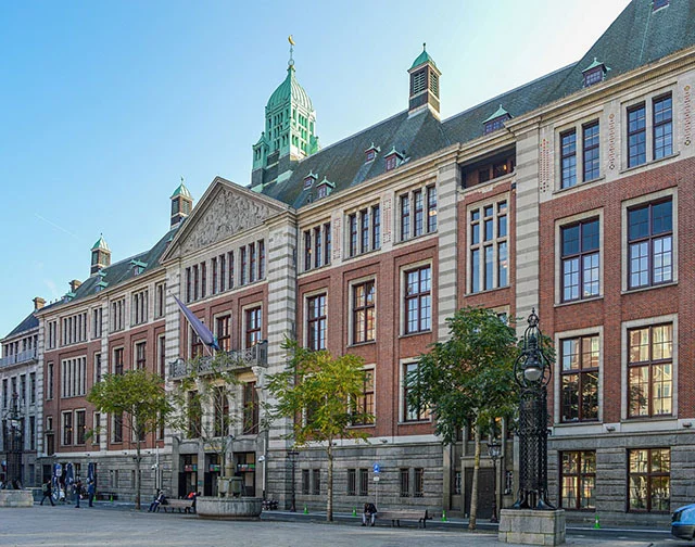 Historic red-brick building with green-roofed tower in a city square. People walk nearby, trees line the front, clear blue sky above.