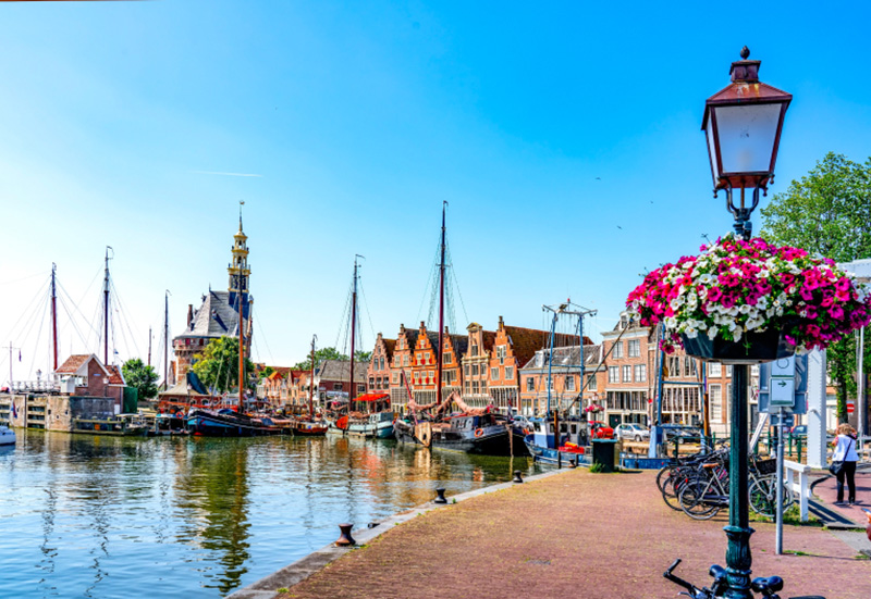Scenic harbor view in Hoorn, Netherlands, featuring historical buildings, moored boats, and colorful flowers on a sunny day. Person stands near bicycles.