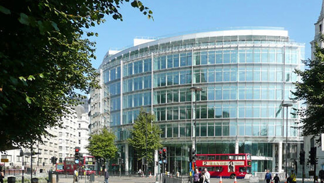 A modern glass building in London, featuring red double-decker buses and several people walking nearby. Trees partially frame the street view.