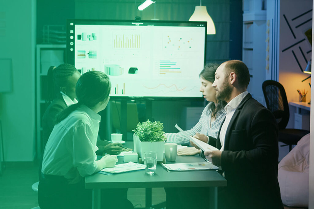 A group of people sit around a table analyzing charts on a screen in a modern office setting, discussing data.